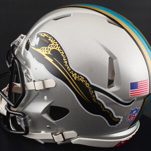 May include: Silver football helmet with black face mask and white chin strap. The helmet features a teal and gold stripe, a black and gold jaguar logo, an American flag, and an NFL logo.