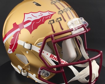 May include: A gold-colored football helmet with a maroon facemask and interior padding. The helmet features a maroon and white spear graphic and several maroon tomahawk graphics. The word "NOLES" is visible on the front of the helmet.
