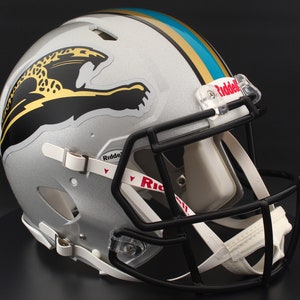 May include: Silver football helmet with a black facemask and white chin strap. The helmet features a jaguar logo in black and gold, with teal and gold stripes on top. The Riddell logo is visible on the facemask and chin strap.