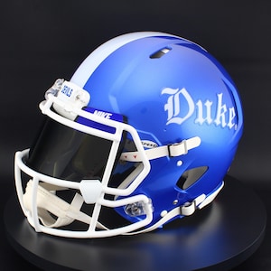 May include: A glossy royal blue football helmet featuring a white center stripe and the word Duke in white lettering on the side. The helmet includes a white face mask, a dark tinted visor, and a small white clip labeled Devils at the top.