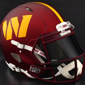 May include: A burgundy football helmet with a yellow stripe and a large yellow "W" logo. The helmet has a dark visor, a burgundy face mask, and white chin strap. The word "Commanders" is visible on the strap.