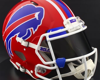 Puede incluir: Un casco de fútbol americano rojo y azul con el logotipo de los Buffalo Bills. El casco tiene una máscara facial blanca, una visera tintada y la palabra "BILLS" impresa en el clip de la visera. Acabado brillante.