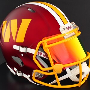 May include: A burgundy football helmet with a yellow and white stripe and a yellow face mask. The helmet features a large yellow "W" logo and a tinted visor. The helmet is a sports collectible.