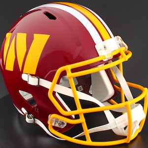 May include: A burgundy football helmet featuring a large yellow letter W on the side. The helmet has a central white and yellow stripe running from front to back, a yellow face mask, and white chin straps attached with clear plastic clips.