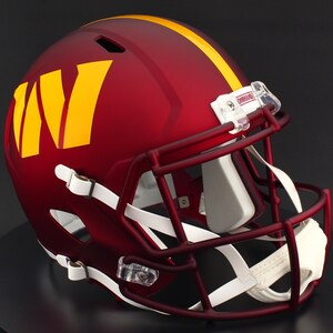 May include: A burgundy football helmet with a yellow stripe and the letter "W" in yellow. The helmet has a maroon facemask, white chin strap, and clear plastic clips. The helmet is on a black surface.