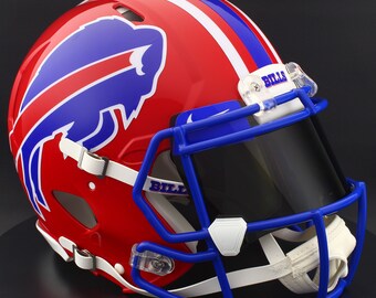 Puede incluir: Un casco de fútbol americano rojo con rayas azules y blancas y el logotipo de los Buffalo Bills. La máscara facial azul y la visera tintada son prominentes. La palabra "BILLS" está impresa en la visera y el casco.