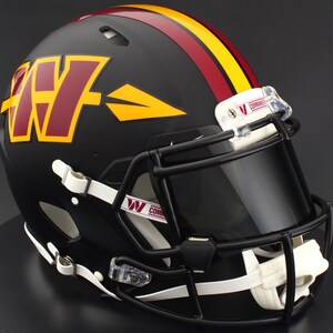 May include: A matte black football helmet featuring a large burgundy and yellow W logo with an arrow design on the side. The helmet has a burgundy and yellow center stripe, a black face mask, a dark tinted visor, and white chin straps with Commanders text.
