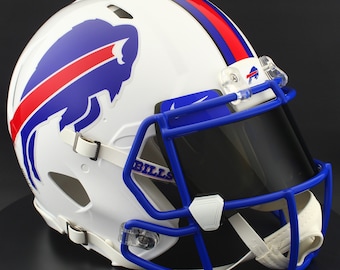 Puede incluir: Casco de fútbol americano blanco con una franja azul y roja y un logotipo de búfalo azul. El casco tiene una máscara facial azul y una visera tintada. La palabra "BILLS" está impresa en el lateral.