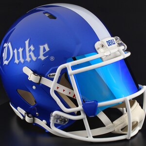 May include: A glossy blue football helmet with a white stripe and face mask. The word "Duke" is displayed in white script on the side. A blue visor and the word "Devils" are visible on the helmet.