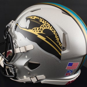 May include: Silver football helmet with a black facemask and white chin strap. The helmet features a jaguar logo in black and gold, with teal and gold stripes. An American flag and NFL logo are visible.
