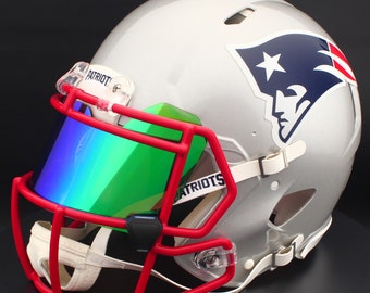 Puede incluir: Casco de fútbol americano plateado con máscara roja y visera multicolor. El casco presenta el logotipo de los Patriots y la palabra "PATRIOTS". Diseñado para la protección durante los partidos de fútbol americano, tiene una correa de barbilla blanca.
