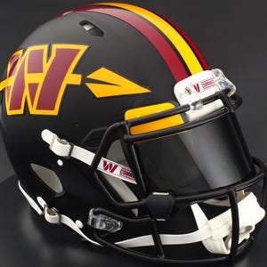 May include: A matte black American football helmet featuring a burgundy and gold W logo with an arrow design on the side. The helmet has a central burgundy and gold stripe, a black face mask, a tinted visor, and white chin straps with Commanders branding.