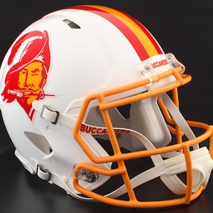May include: White football helmet with orange face mask and stripes. The helmet features a team logo of a pirate with a red hat and the word "BUCCANEERS" in red. The helmet has a clear visor and white chin strap.