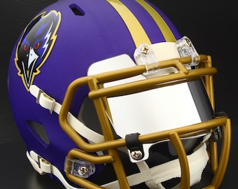 May include: A purple football helmet with gold stripes and a gold facemask. The helmet features a team logo on the side and a clear visor. The helmet is designed for protection during American football games.