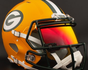 May include: A glossy, gold-colored football helmet with a green and white stripe and the letter "G" on the side. The helmet has a dark green face mask and a red-tinted visor. White chin strap and clear clips secure the helmet.