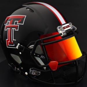 Puede incluir: Un casco de fútbol americano negro con una franja roja y blanca y una visera roja. El casco presenta un gran logotipo "T" rojo y blanco y las palabras "Red Raiders" en el clip de la visera.