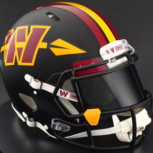 May include: A matte black football helmet featuring a large burgundy and yellow W logo with an arrow design on the side. A yellow and burgundy stripe runs down the center. The helmet includes a black face mask, a dark visor, and white chin straps.