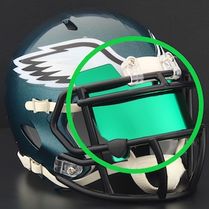 May include: A teal and white football helmet with a green visor. The helmet has a black facemask and white straps. The team logo is a white eagle head with black accents.