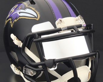 May include: A black football helmet with a black facemask, featuring the Baltimore Ravens logo on the side. The helmet has purple stripes and white and clear straps. The visor is clear.