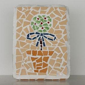 May include: A mosaic tile artwork depicting a potted plant with a green leafy top, red berries, a brown stem, and a blue bow. The pot is brown and the background is beige.