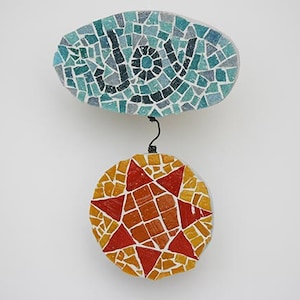 May include: A mosaic wall hanging with the word "Joy" in blue and black tiles on a light blue background. A red star with orange and yellow tiles hangs below the word.