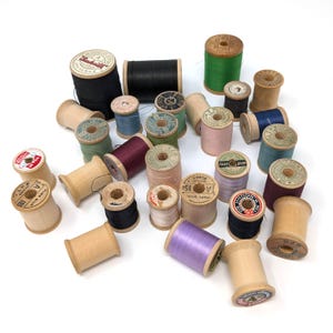May include: A collection of vintage wooden sewing thread spools in various sizes and colors, including black, green, blue, purple, pink, and natural wood tones. Many spools feature paper labels with printed brand names and yardage details.