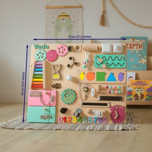 May include: A wooden activity board for children with a variety of textures and activities, including a clock, gears, a lock, a chain, a slider, a zipper, a door, a bead maze, and a xylophone. The board is painted in bright colors and has the name "Veda" written on it. The board measures 27.5 inches wide and 23.6 inches tall.