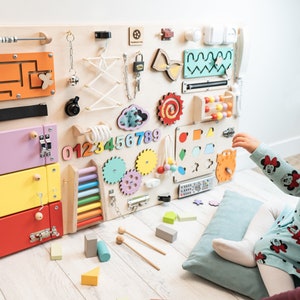 May include: A wooden activity board with various textures and activities for children. It features a maze, a lock and key, a sliding door, a gear system, a bead roller, a xylophone, and a number puzzle. The board is mounted on a wall and a child is interacting with it.