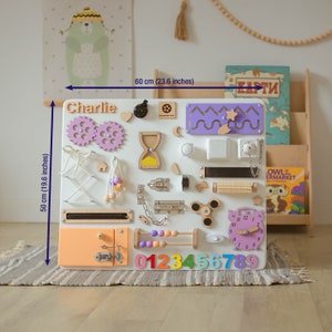 May include: A white wooden activity board with various textures and activities for children. It features a clock, a sliding door, a chain, a latch, a spinning wheel, a bead maze, a shape sorter, a zipper, a toggle, a knob, a mirror, a bell, a gear, a  labyrinth, and a number line. The board is labeled "Charlie" and measures 23.6 inches by 19.6 inches.