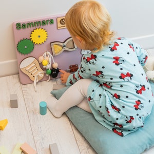 May include: A pink wooden activity board with the name "Sammara" written on it. The board has various activities for children, including a lacing shoe, a spinning gear, a bead maze, and a sliding latch. A young child is playing with the board.