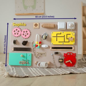 May include: A beige wooden activity board with various textures and activities for children. It features a clock, a chain, a lock, a maze, a door with a latch, a roller, gears, a switch, and a knob. The board is labeled "Sophia" and measures 15.7 inches by 23.6 inches.