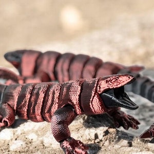 May include: Two red and black articulated toy lizards with a segmented body and a textured surface. The lizards are posed on a rocky surface.