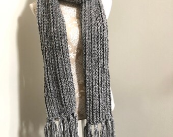 May include: A gray knitted scarf with fringe on the ends. The scarf is displayed on a white mannequin.