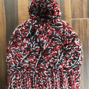 May include: A red, black, and gray knitted beanie with a fluffy pom pom on top.