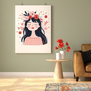May include: A framed illustration of a woman with closed eyes wearing a floral crown. The woman has long black hair and is wearing a pink sweater with white polka dots. The background is a light green wall with a small table and a vase of red flowers.