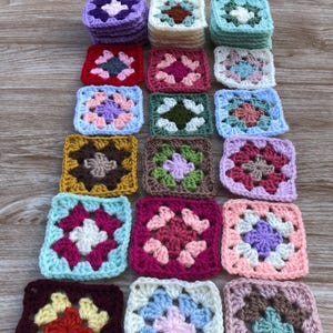 May include: A collection of multi-colored crochet granny squares arranged in a pyramid formation. Each square features a central flower-like pattern in various color combinations including purple, red, pink, green, blue, yellow, and white yarn.