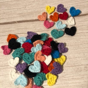 Può includere: Una collezione di piccoli cuori all'uncinetto in vari colori, tra cui rosa, giallo, blu e nero. I cuori sono sparsi su una superficie di legno di colore chiaro. I cuori fatti a mano sono un oggetto artigianale.