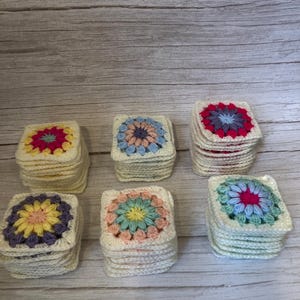 Pode incluir: Seis pilhas de bases de copos quadradas de crochê com desenhos florais. Cada base de copo apresenta um esquema de cores diferente, incluindo rosa, amarelo, azul e roxo, em um fundo creme. As bases de copos são dispostas em uma superfície clara com grão de madeira.