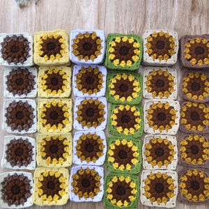 Pode incluir: Uma coleção de trinta motivos quadrados de crochê dispostos em uma grade. Cada motivo apresenta um design central de girassol marrom com pétalas e bordas amarelas, brancas, verdes ou marrons, exibindo texturas intrincadas de fio.