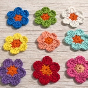 Może przedstawiać: Dziewięć kolorowych, szydełkowych kwiatów z pomarańczowymi środkami. Kwiaty są w różnych kolorach, w tym niebieskim, zielonym, białym, żółtym, różowym, fioletowym i czerwonym. Ułożone na jasnej powierzchni imitującej drewno.