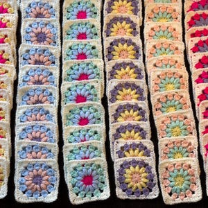 Può includere: File di colorati quadrati granny all'uncinetto in varie tonalità pastello, tra cui rosa, blu, giallo e viola. Ogni quadrato presenta un motivo floreale con un bordo bianco. I quadrati sono disposti in colonne ordinate, mostrando l'artigianato fatto a mano.