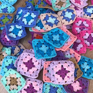 Pode incluir: Uma coleção de quadrados de crochê granny coloridos em vários tons de azul, roxo, rosa e verde. Cada quadrado apresenta um design floral central, criando um efeito de patchwork. Os quadrados estão espalhados em uma superfície de madeira.