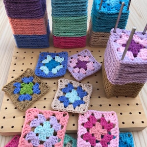 Puede incluir: Una colección de cuadrados y pilas de ganchillo coloridos hechos a mano. Los cuadrados presentan patrones florales en varios colores, incluyendo rosa, azul, morado y verde. Algunos cuadrados están apilados sobre bases de madera con varillas metálicas.