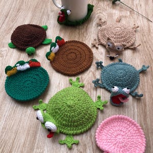 Op de afbeelding: Een verzameling gehaakte onderzetters met diverse dierontwerpen, waaronder een bruine schildpad, een groene kikker, een roze slak, een beige egel en een blauw wezen met een rode mond, naast een witte keramische mok met een groene kikker.