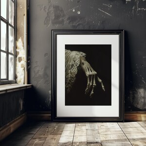 K&ouml;nnte beinhalten: Ein schwarzer Bilderrahmen mit einem wei&szlig;en Passepartout. Das Bild zeigt eine skelettartige Hand, die aus einem wei&szlig;en Stoff herausragt. Der Hintergrund ist dunkel.