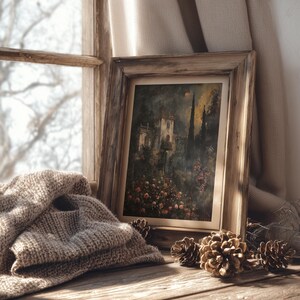 May include: A framed landscape painting with a rustic wooden frame rests on a wooden surface, next to a folded beige knit blanket and several pine cones. The painting depicts a building surrounded by trees and flowers. The text "DARKENDEDBRUSH" is visible.
