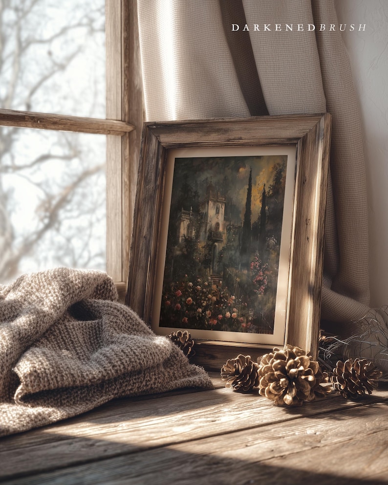 May include: A framed landscape painting with a rustic wooden frame rests on a wooden surface, next to a folded beige knit blanket and several pine cones. The painting depicts a building surrounded by trees and flowers. The text "DARKENDEDBRUSH" is visible.