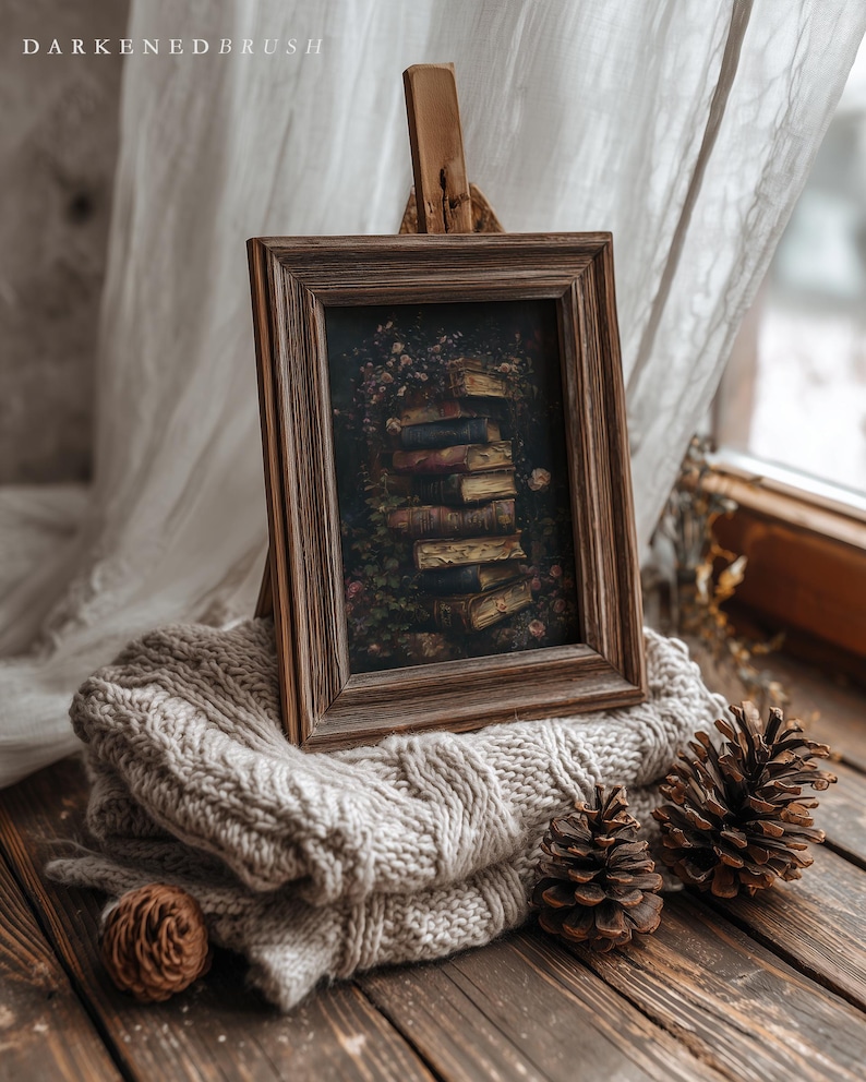 May include: A framed artwork featuring a stack of books, displayed on a wooden easel. The artwork is set on a gray knitted blanket, with pine cones on a wooden surface. The frame is brown, and the artwork has a dark, vintage aesthetic.