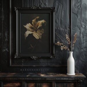 May include: A black framed picture with a dried white lily flower in a dark background. The picture is hanging on a black wall with a white vase and dried flowers on a dark wooden table.