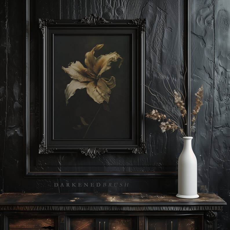 May include: A black framed picture with a dried white lily flower in a dark background. The picture is hanging on a black wall with a white vase and dried flowers on a dark wooden table.